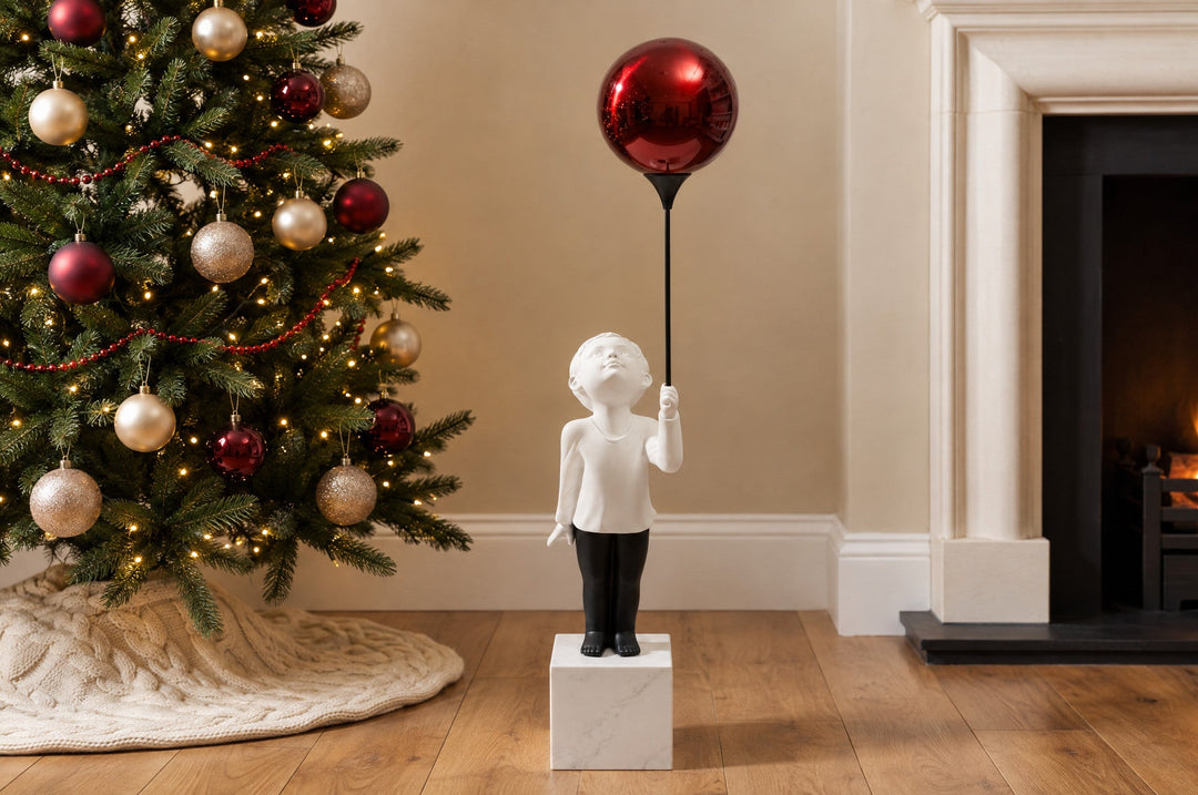 AeroCherub, a boy sculpture holding a red balloon, stands in a neutral living room, and the polished red reflects the Christmas tree and the firelight.