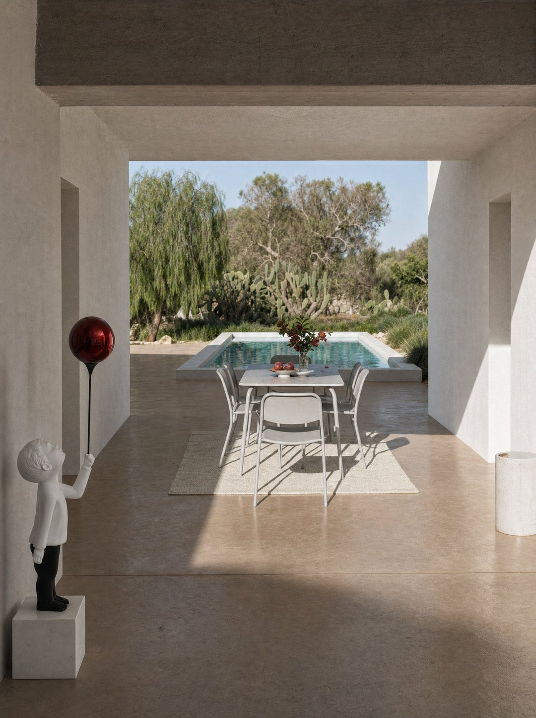 A hallway threshold looking out to a swimming pool and wild garden with cacti and olive trees beyond, an AeroCherub sculpture on a small pedestal in the left foreground, partly cut by the wall edge, facing the garden.