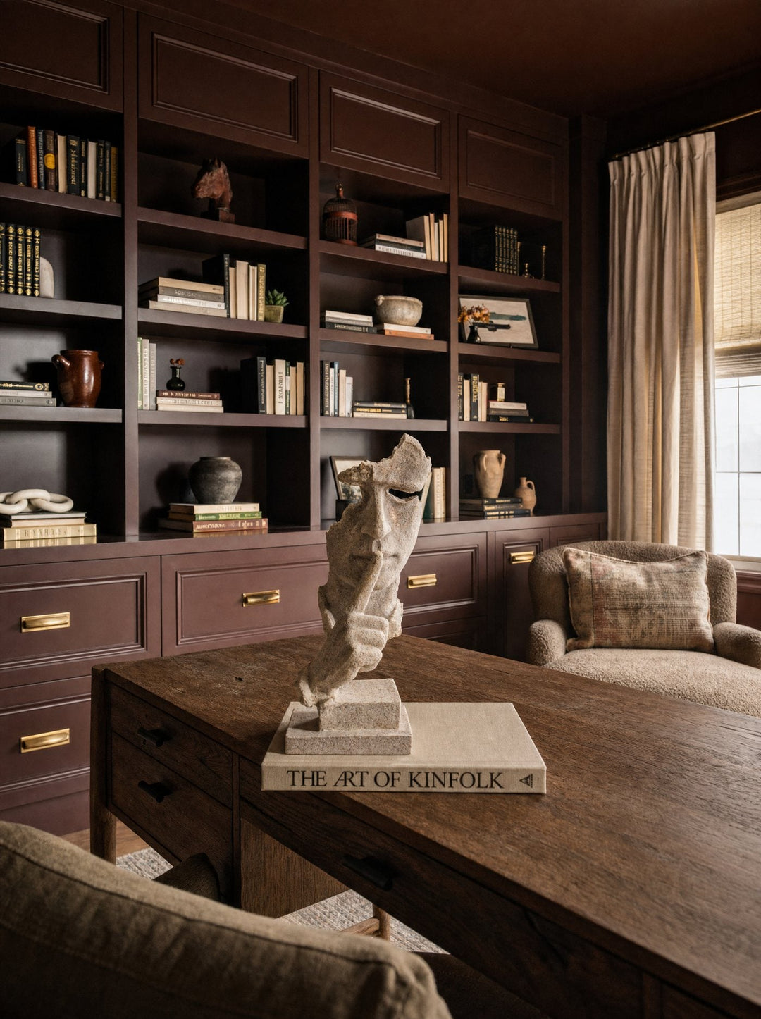 A carved stone face sculpture with a finger to its lips, resting on a book titled The Art of Kinfolk, on a wood desk in front of a dark built-in library.