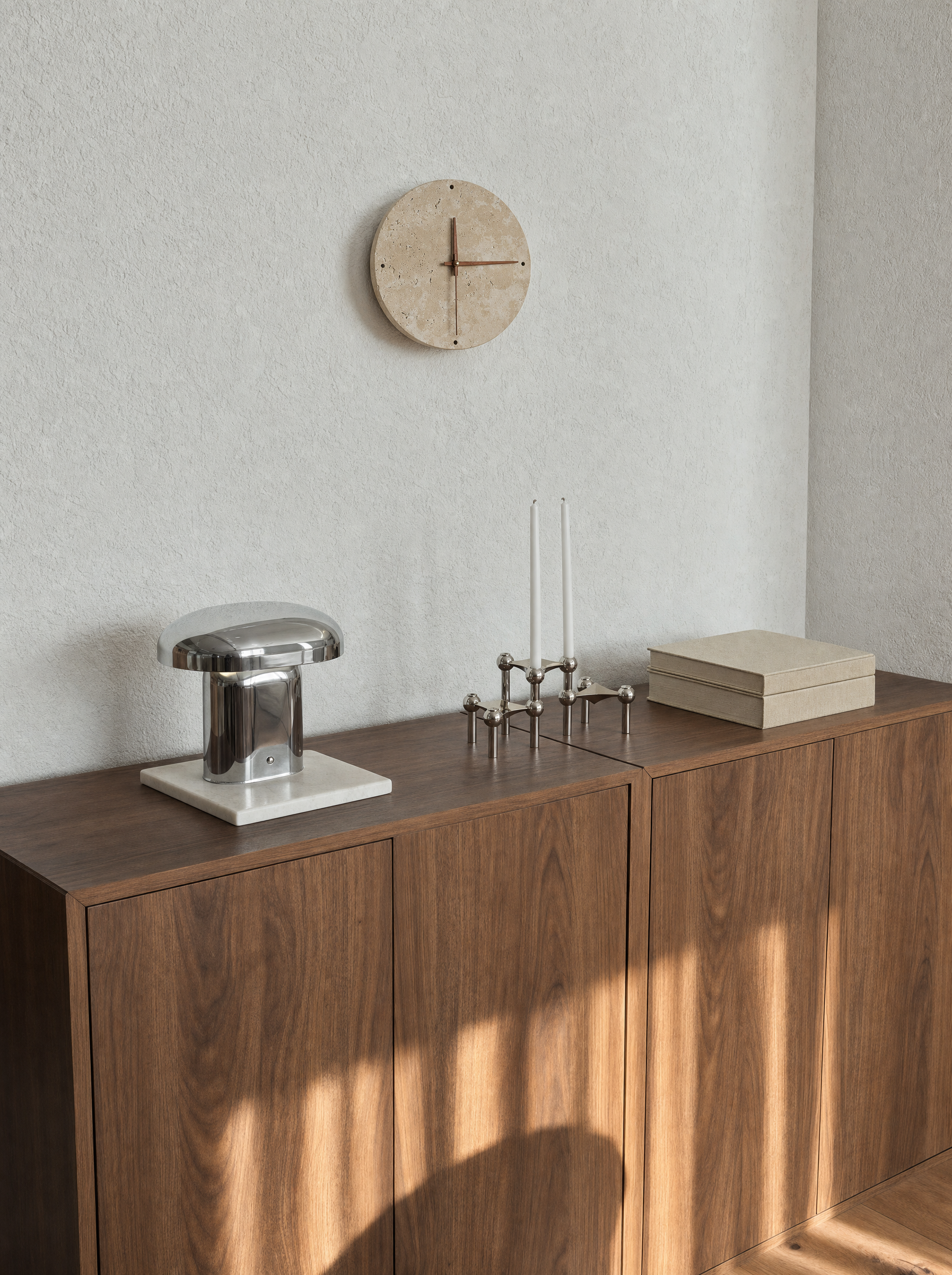 Adagio travertine wall clock, light off, above a walnut console with a chrome mushroom lamp and candle holders