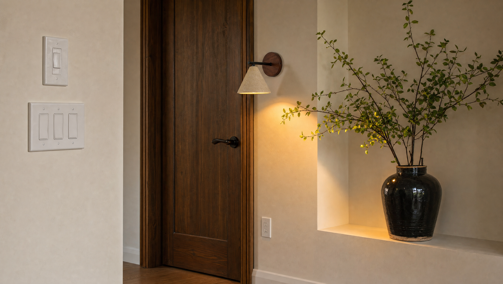 Fonte wall lamp beside a wooden door, warm light on green branches in a plaster niche