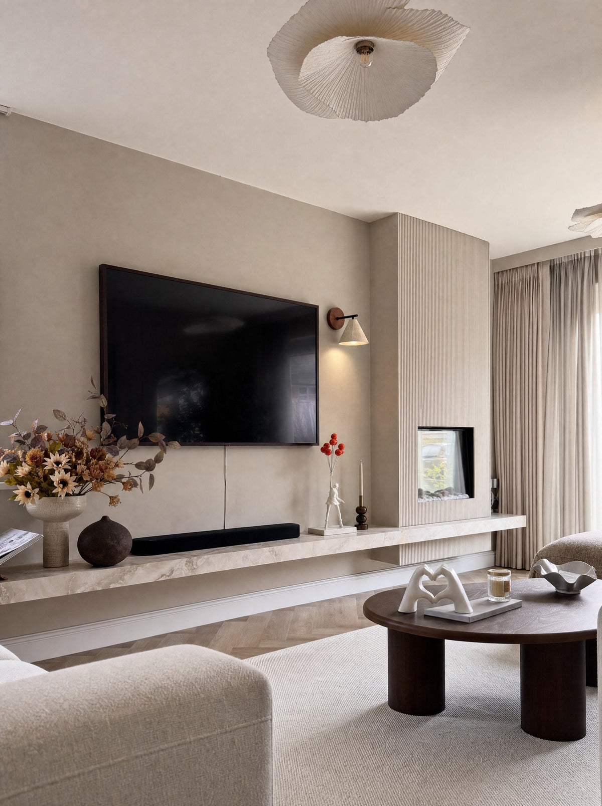 Elevate, a white figure in mid-stride with eight red balloons pulling it up, its base of white marble on a long floating shelf of beige stone beside a black soundbar, to one side of a dark television, with Fonte, a wall lamp, its cone shade of stone on a black arm from a round walnut disc, switched on above it on the same wall, and AmorSignum, two hands forming a heart on two separate oval feet, on a book on a round dark wood coffee table, in a living room with a slatted fireplace column