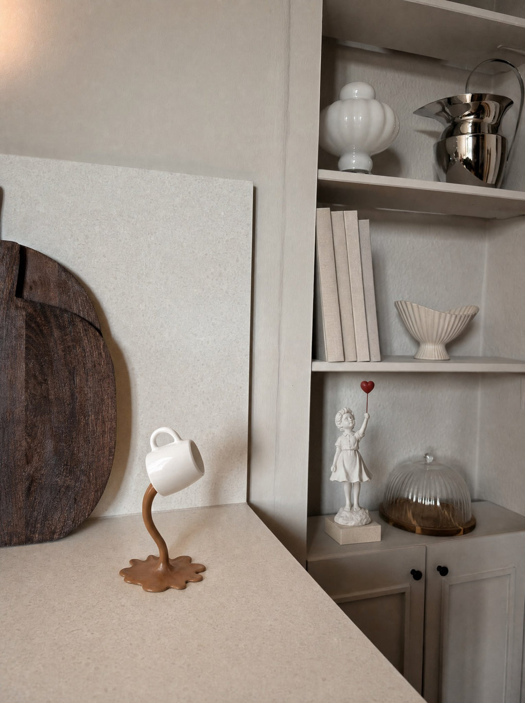 CaffeInAria, a white cup held tilted at the top of a curved coffee pour in matte resin that rises out of a splashed pool, standing on a cream terrazzo kitchen counter near a dark wood cutting board, with Ascend, a small figure in white stretching up toward a red heart held on a thin rod, on its marble base on the cabinet top inside a cream recessed shelf, among white china and a glass cloche