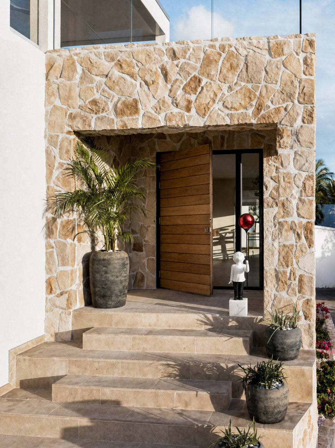 AeroCherub, a tall white cherub with its face turned up to a red sphere of polished chrome, its white marble block set on the paving of a stone-clad entrance beside an open slatted wood door, with a potted palm to the left, stone steps and pink bougainvillea in the sun