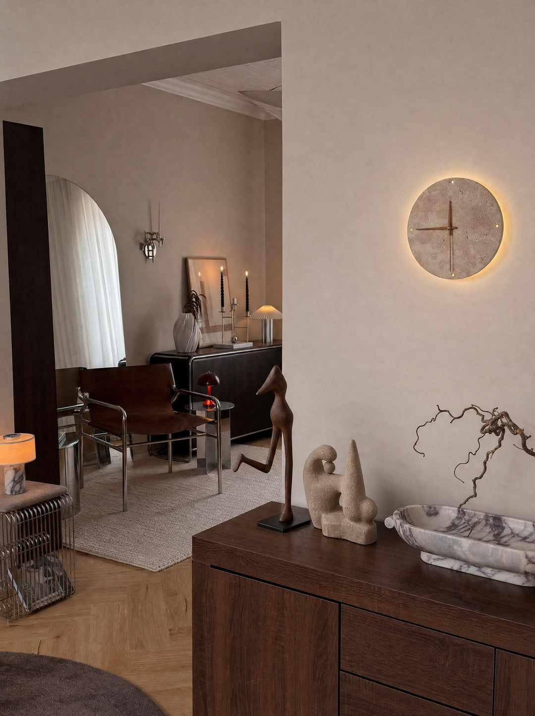 Adagio, a wall clock in a solid disc of travertine stone with three walnut-tone wooden hands and four small holes at twelve, three, six and nine, no numbers, lit with a warm halo on the hallway wall around its rim, with Archetypes, two figures, the tall one slender and the short one solid, on a walnut cabinet beside an oval bowl of purple stone holding a dry branch, and a doorway view of a leather and chrome chair in the room beyond, on a herringbone wood floor