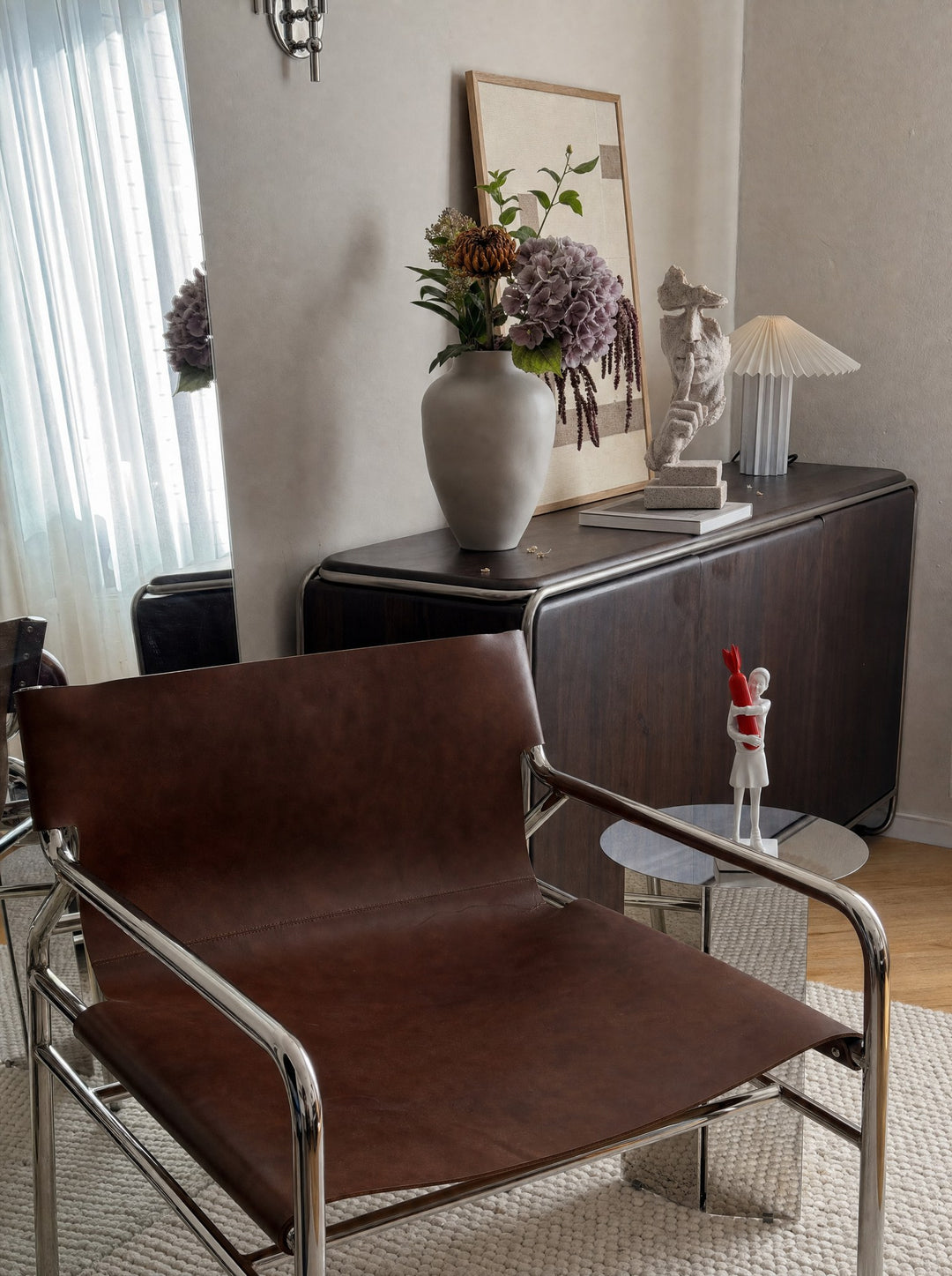 Disarm, a figure in white with both arms wrapped around a red bomb, on a round chrome side table next to a brown leather chair on a tubular chrome frame, with SilentMuse, a blank-faced figure, one finger raised against its lips, standing on a rounded dark wood cabinet with chrome trim between a leaning framed painting and a small white pleated lamp, near a white vase of purple hydrangeas and a cream wool rug