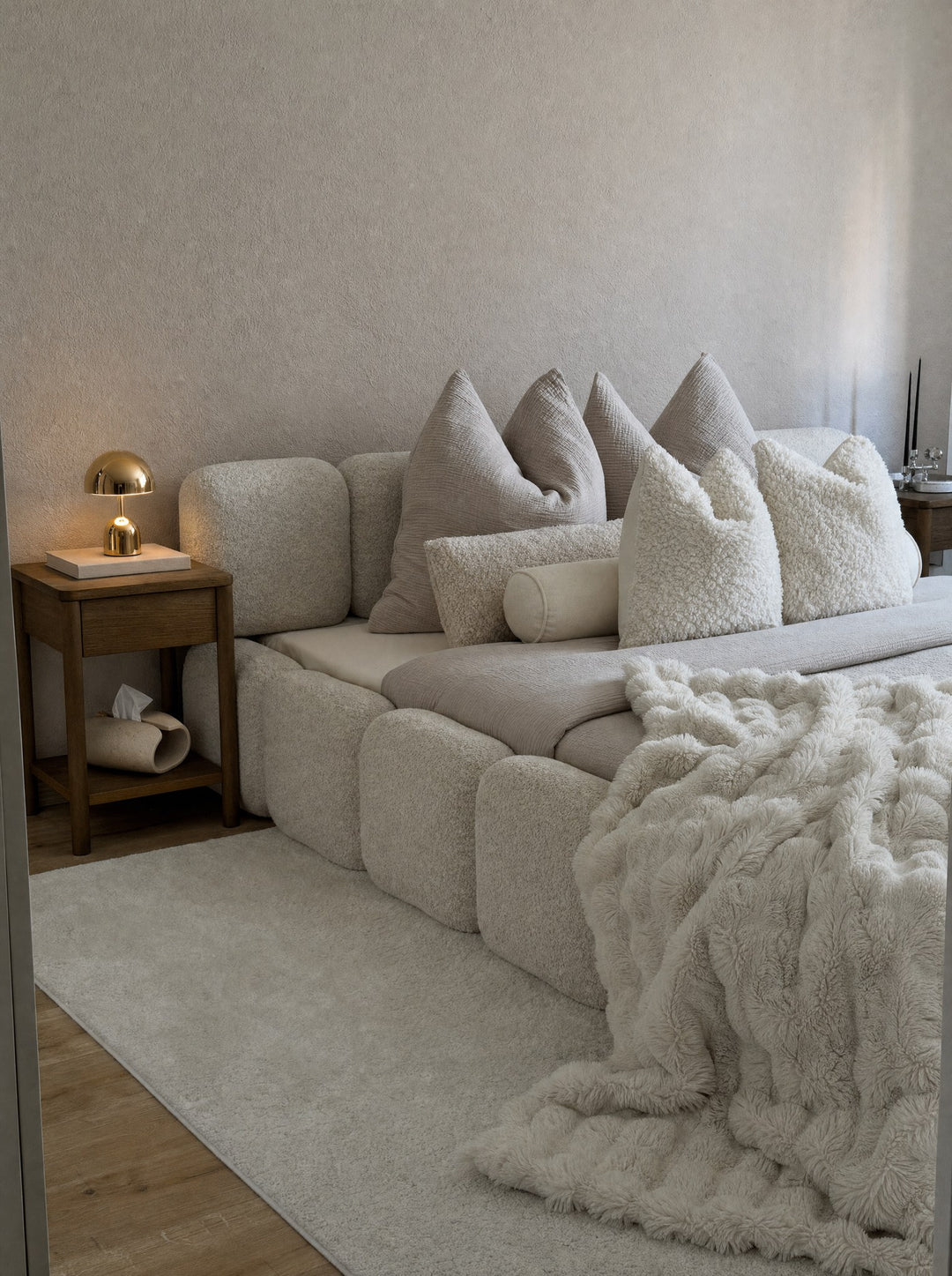 Vespers, a gold cordless lamp, its mushroom dome lit above two books on an oak nightstand, with Volute, a tissue box in matte ceramic, one band curled into a single loop with a tissue drawn part way out, on the shelf below, beside a bed in cream boucle with a thick throw, against a cream plaster wall in afternoon light