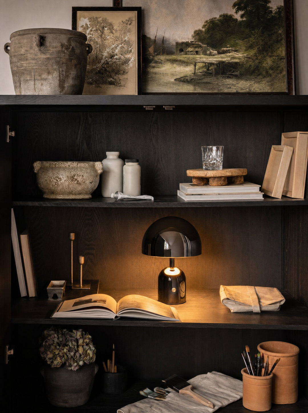 Vespers, a black cordless lamp, its round dome switched on, standing on a dark wood shelf inside an open cabinet behind an open book, beside a folded linen roll and a pair of gold-tone candlesticks, with stoneware pots and two small paintings on top of the cabinet and terracotta pots of brushes on the shelf below