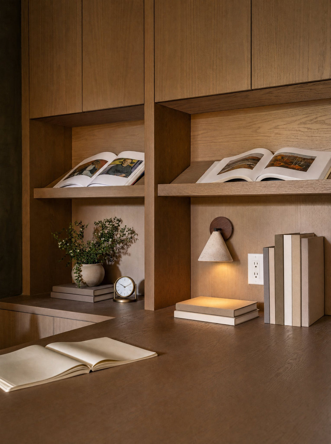 Fonte, the wall lamp, switched on against the oak back panel of a built-in shelf, its stone cone hanging from a walnut disc and its light falling onto a pile of two books beside books standing on end, with open art books on sloped shelves above, a small potted plant to the left and an open notebook on the desk in front