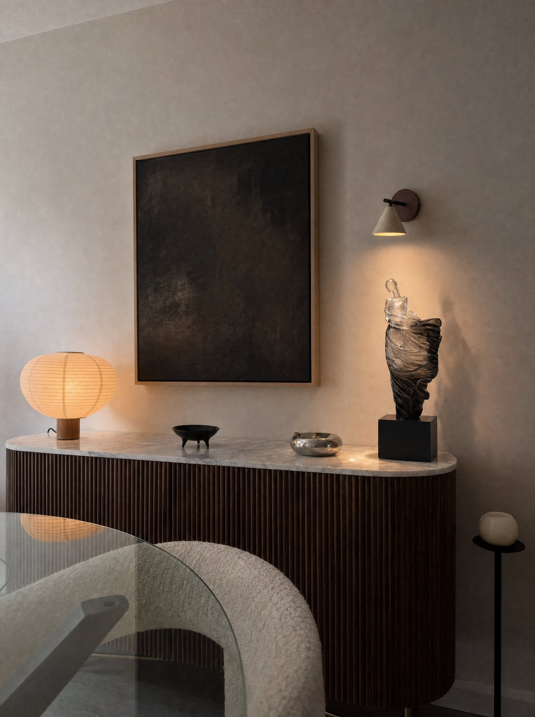 Dualité, a figure in translucent resin, pale near the head and darkening toward the foot, on a black plinth on the marble top of a fluted dark wood sideboard, lit from above by Fonte, the stone cone wall lamp on its dark walnut disc on a plaster wall, beside a large dark framed painting, with a bouclé chair and a glass table in front