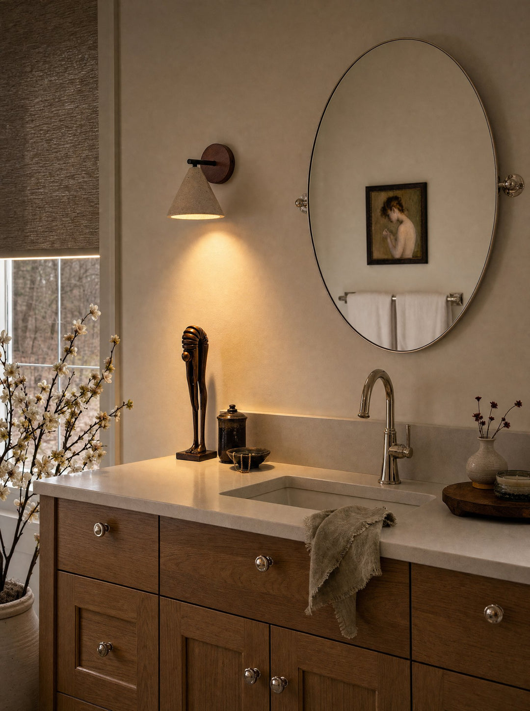 Fonte, a stone and walnut wall lamp on a short black arm, lit beside an oval bathroom mirror, its light falling down the wall toward The Echo, a little figure of thick stacked discs on two straight legs, reddish-brown in patina, standing on a cream stone counter above an oak vanity, with white blossom branches by the window and a linen towel at the sink