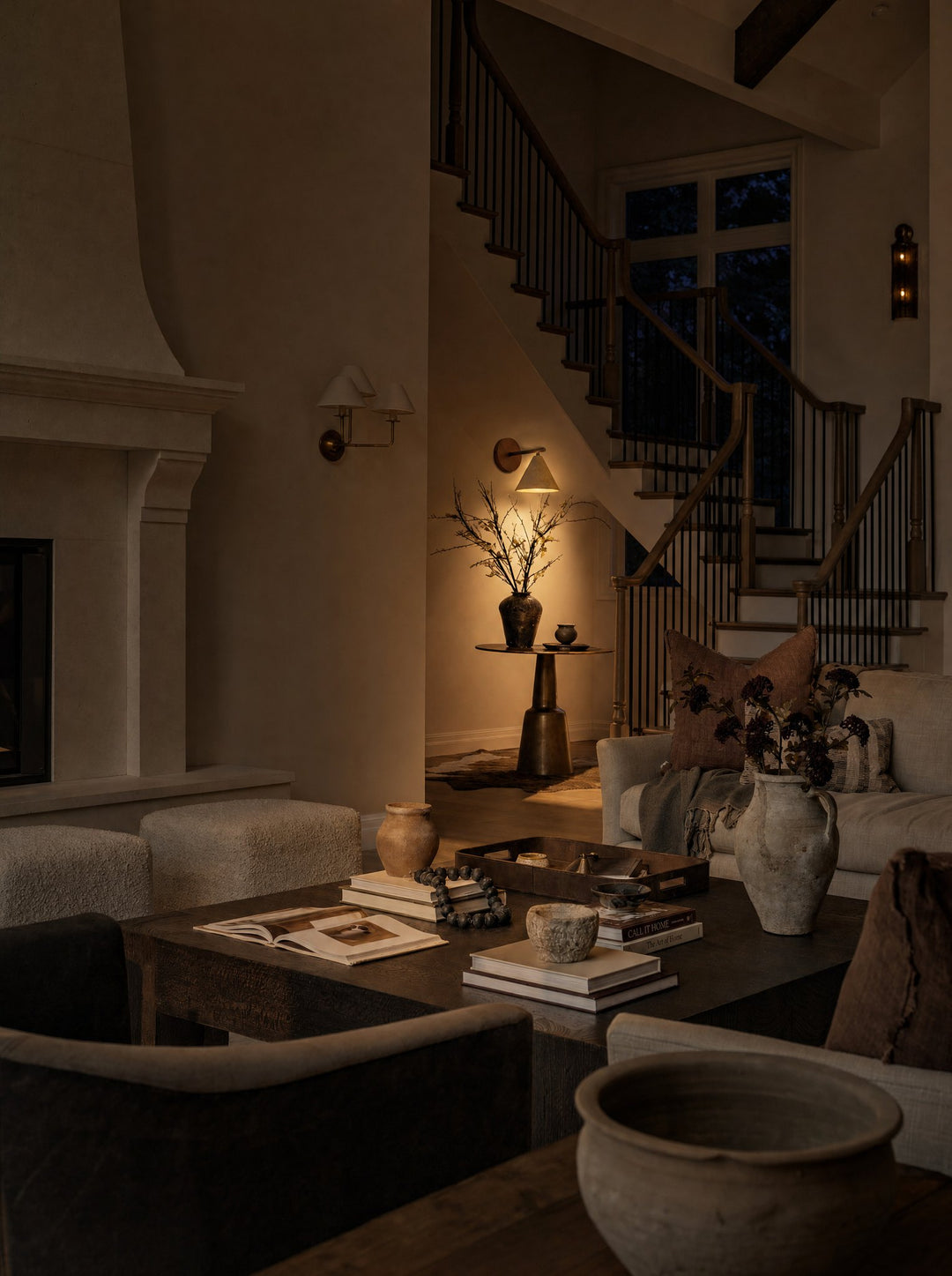 Fonte, the cone lamp of stone on walnut, lit by the bottom of the staircase at the rear of a dim living room in the evening, its light falling on a dark vase of bare branches on a slim pedestal table, with a stone fireplace, a low wood table of books and clay vessels, and a cream sofa piled with brown cushions