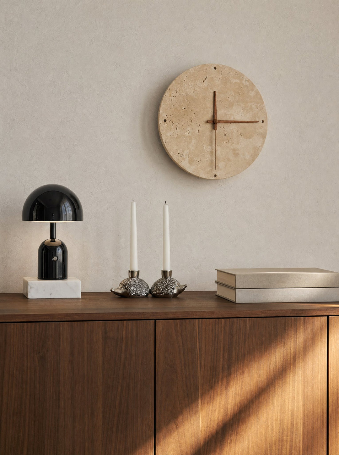 Adagio, a round wall clock of pitted travertine stone, its hands of walnut-tone wood and a small hole at the top, bottom and each side of the face, its light off, above a walnut sideboard that holds Vespers, a black mushroom lamp on a block of white marble, a pair of white candles in silver hedgehog holders and two cream books, with sun falling in stripes across the cabinet doors