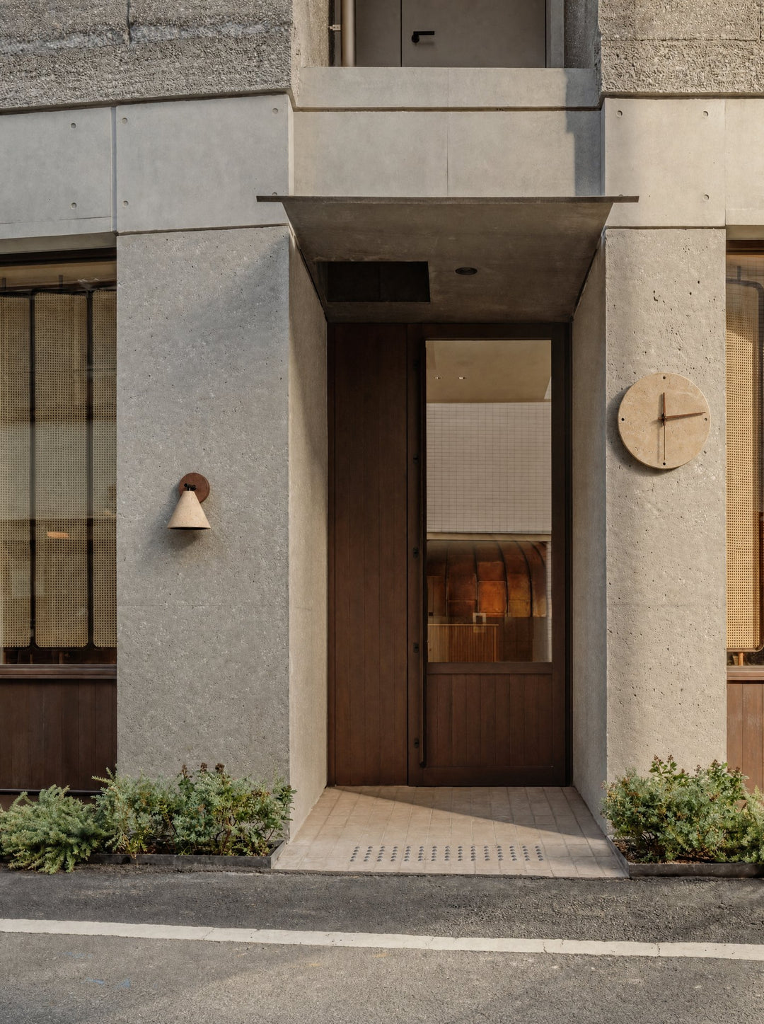 Fonte, a stone cone lamp below a round walnut plate, mounted on the left pillar of a concrete entrance, and Adagio, a round clock of pale stone with dark wooden hands, on the right pillar, both switched off in daylight on either side of a tall wooden door under a concrete canopy, with low shrubs along the pavement