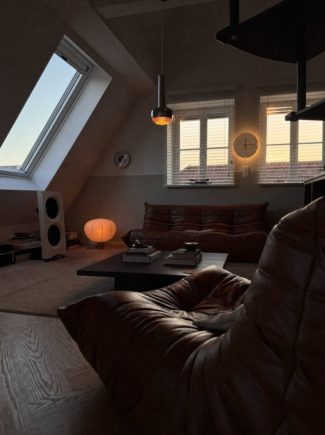 Adagio, a travertine clock with slim wooden hands, its warm halo spread on the wall around the rim, hung between two windows under the sloped ceiling of an attic room at dusk, with a skylight, a low leather sofa, a leather lounge chair close in front and a dark coffee table holding piles of books, on a pale wood floor