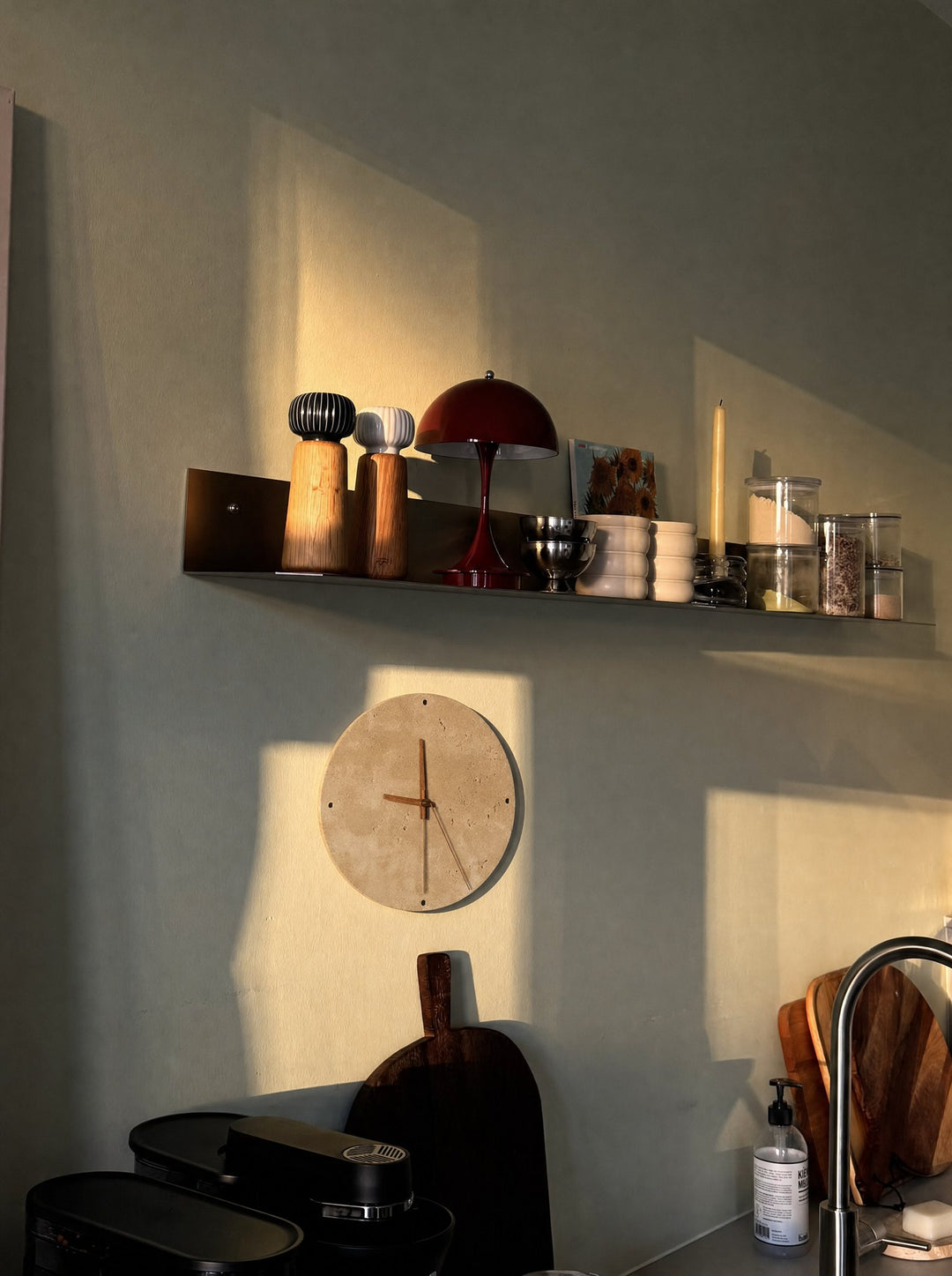 Adagio, the travertine clock, a round stone face with wooden hands in a walnut tone and no numbers, its light switched off, in a patch of sun on a gray-green kitchen wall, below a long metal shelf of spice grinders, stacked bowls, a candle and glass jars, above dark wood cutting boards