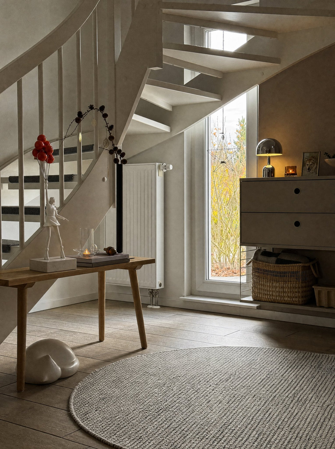 Vespers, a silver mushroom lamp, lit and standing on a pale two-drawer cabinet beside a tall window in a white stair hall, with Elevate, a striding figure in white pulled along by its eight red balloons, set on white marble atop a wooden bench across the hall beside a lit tea light, above a round gray woven rug on a wood floor