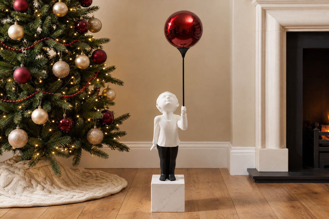 AeroCherub, a boy sculpture holding a red balloon, stands in a neutral living room, and the polished red reflects the Christmas tree and the firelight.