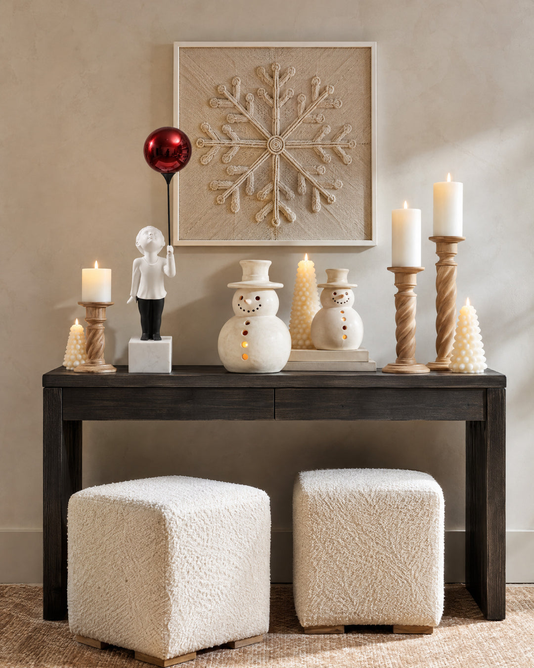 On a console of dark wood against a beige wall, AeroCherub stands among candles and a white porcelain snowman, below a hanging yarn snowflake.