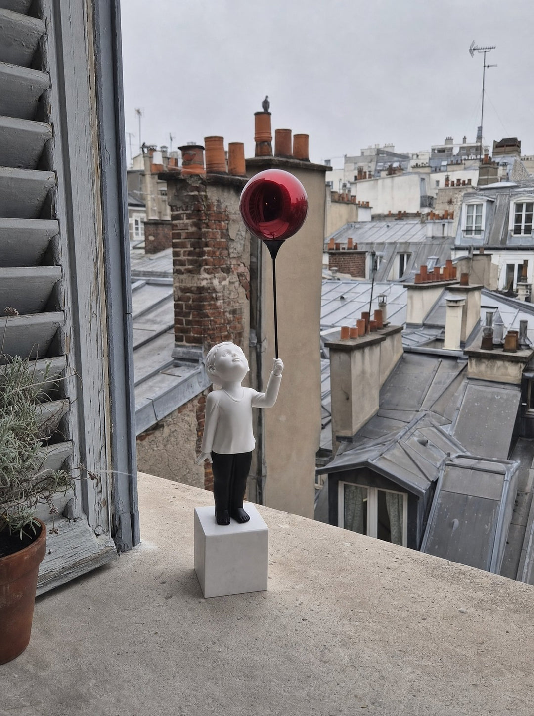 AeroCherub, the boy, a matte white resin figure in black trousers, on a white marble cube set on a stone windowsill, looking up at a red mirror-polished metal balloon. Beside him, a clay pot of green leaves and a weathered blue shutter. Below, gray zinc Paris rooftops and brick chimneys, a pigeon resting on one, under an overcast sky.