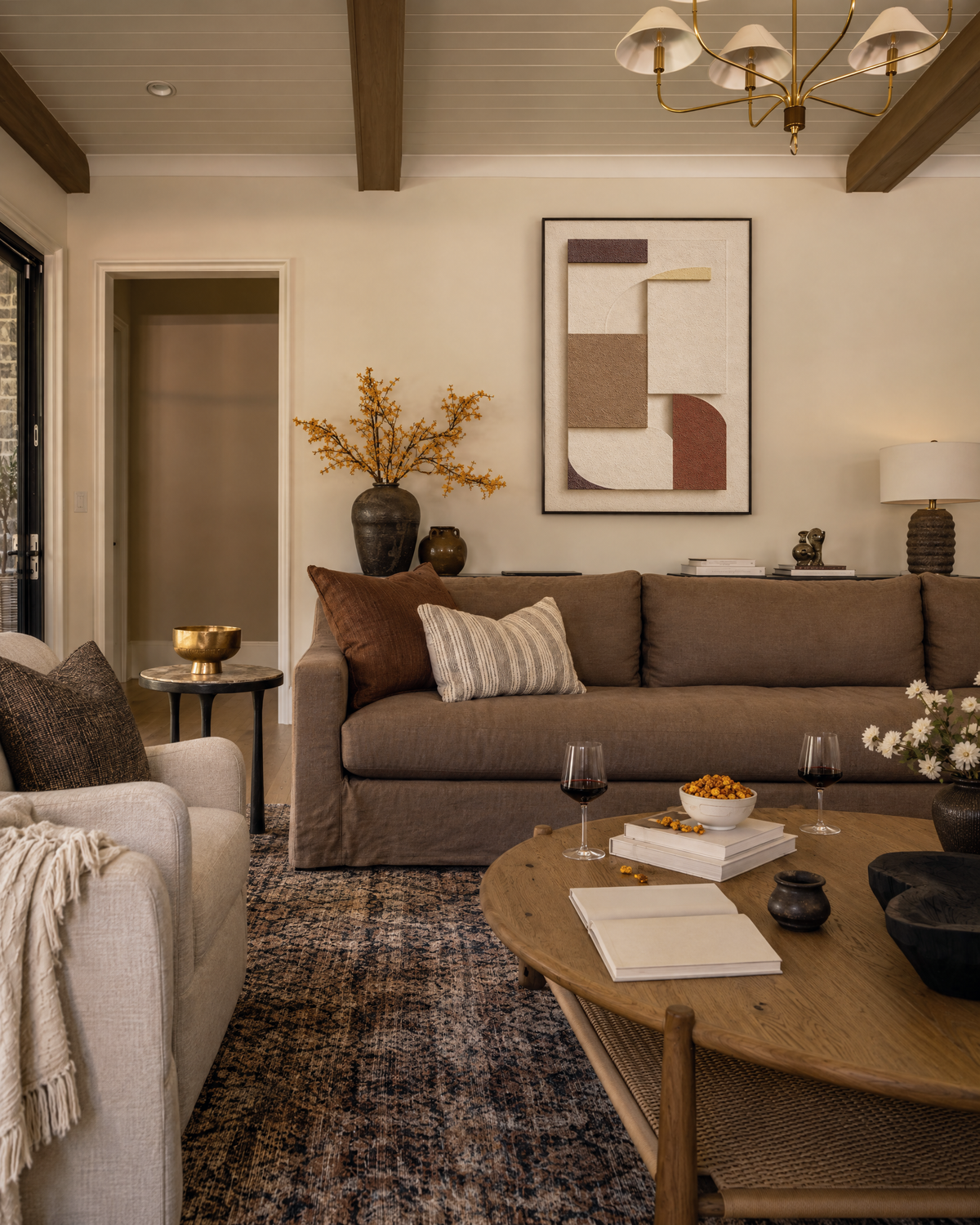 A warm living room seen as a whole, with Rilievo in Terra in a slim black frame above a long brown sofa, under dark ceiling beams and a chandelier, with a round coffee table in front