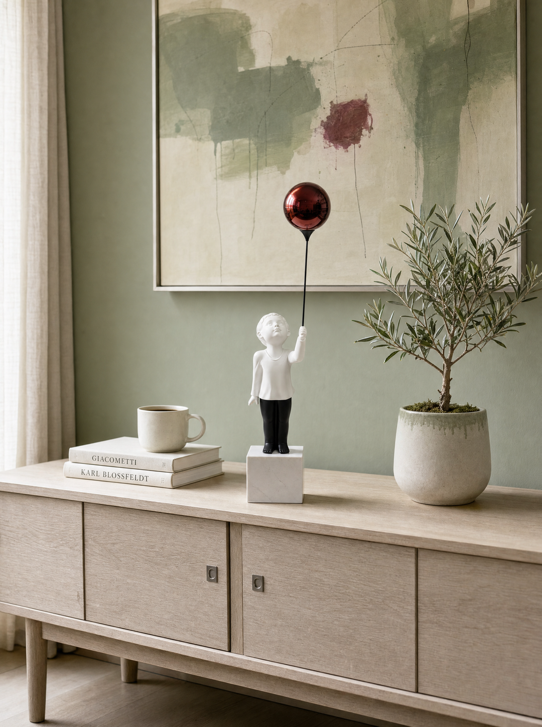 AeroCherub Sacred in the Mundane — sculpture on bleached oak mid-century credenza against sage wall with art books, cream mug, and olive topiary, environmental view