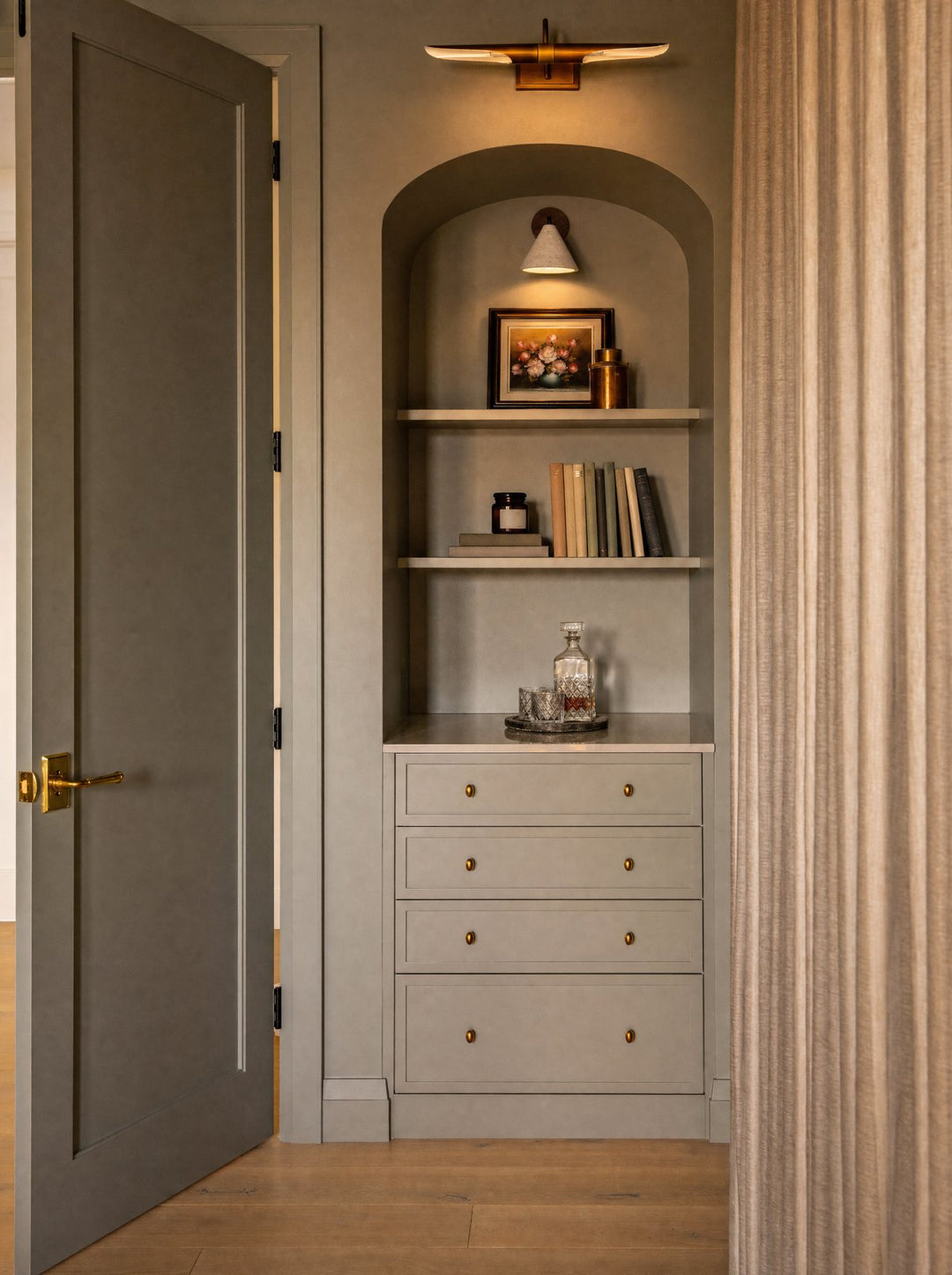 Fonte, a wall lamp with a stone shade and a walnut disc, mounted inside an arched alcove above a shelf of books, seen through an open gray-green door