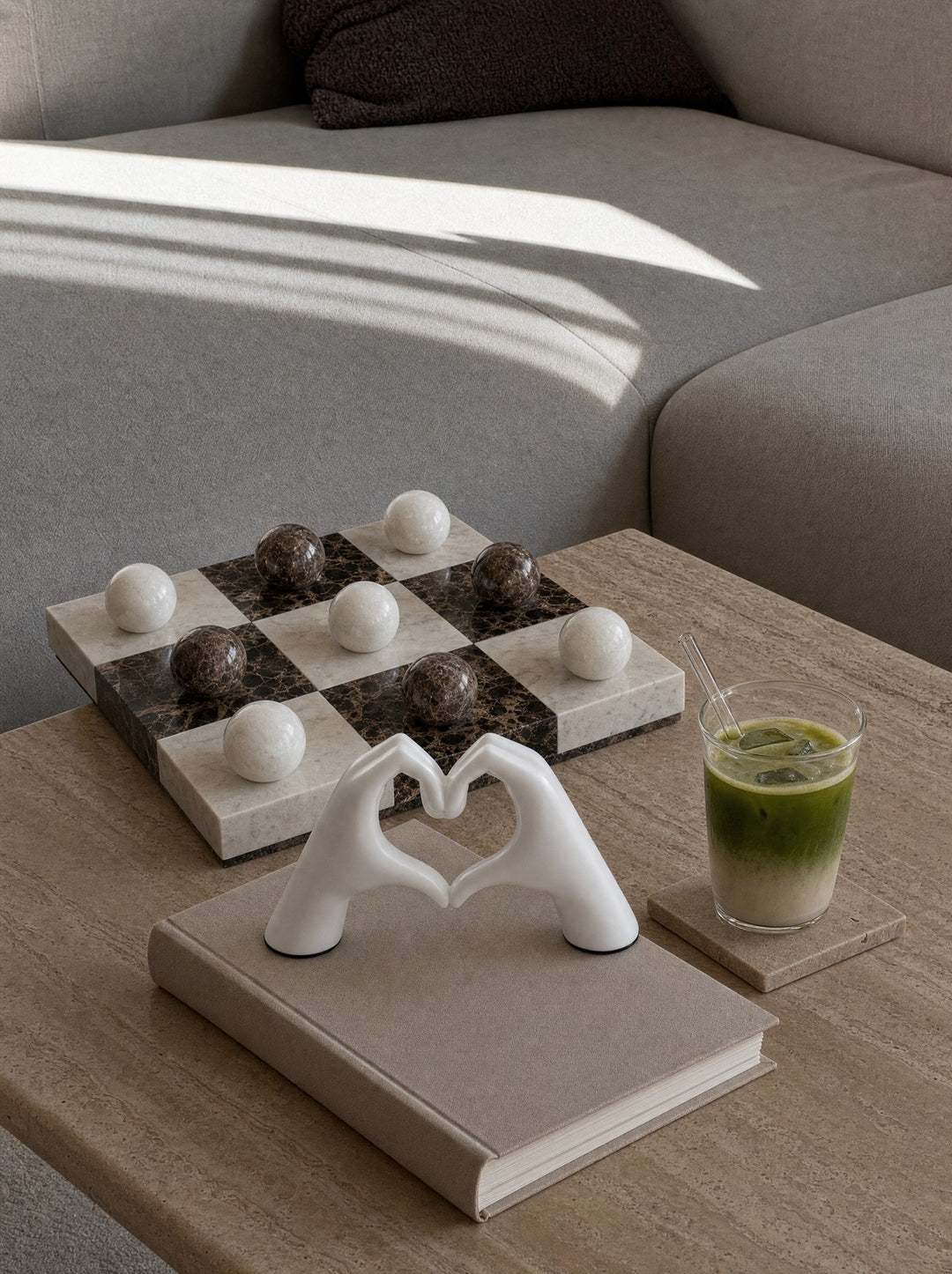 Lapilli, a marble tic-tac-toe board with nine stone spheres, five white and four brown, on a travertine coffee table, with AmorSignum, two white hands with fingertips meeting to form a heart, standing on a hardcover book at the front edge beside a glass of iced matcha on a stone coaster, and slanting afternoon sun across the gray sofa behind