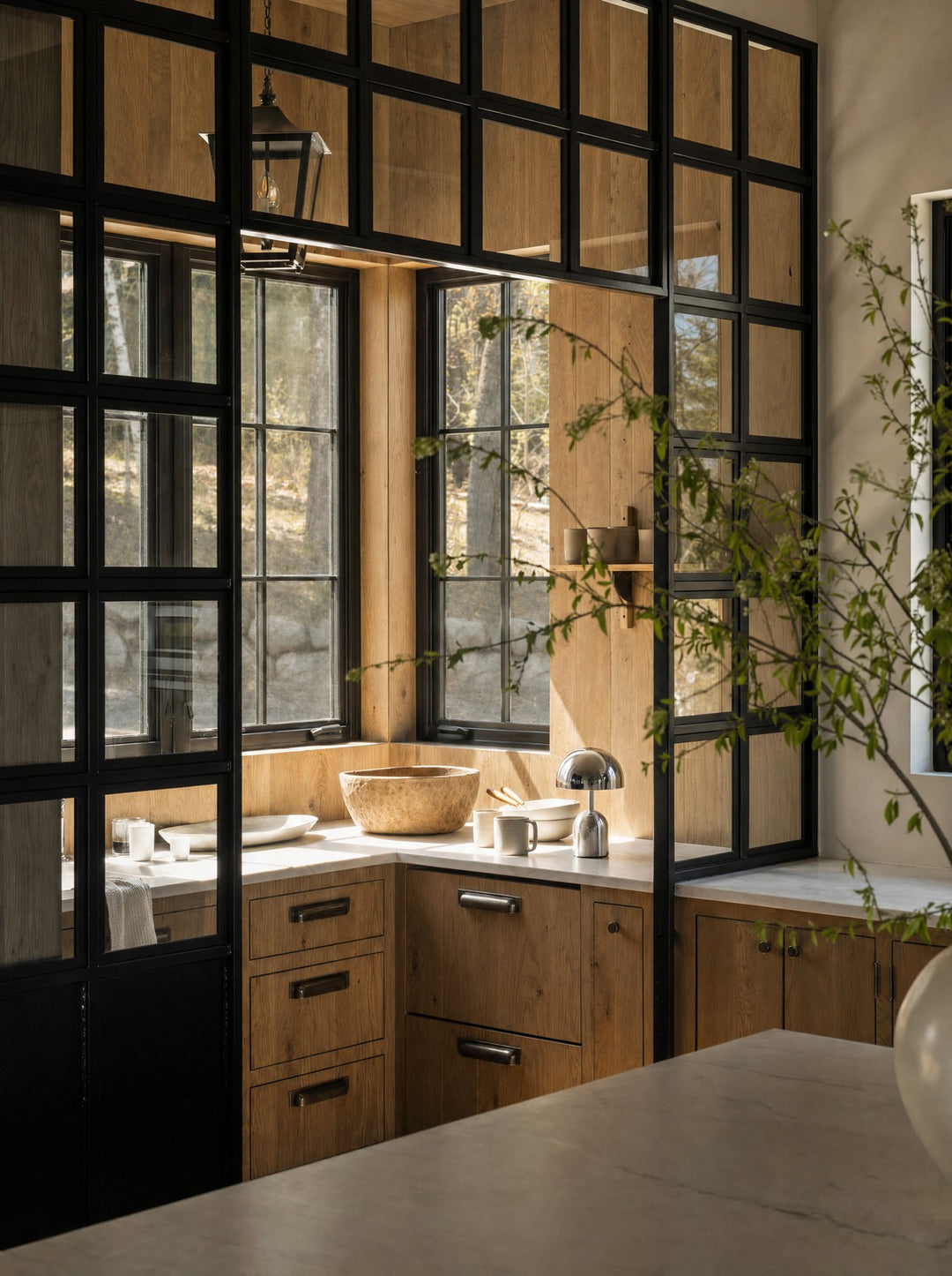 Vespers, a cordless silver mushroom lamp, unlit on the counter beside a window in afternoon light, with a large stone bowl and a ceramic cup nearby, in a kitchen of oak cabinets behind a black steel and glass partition