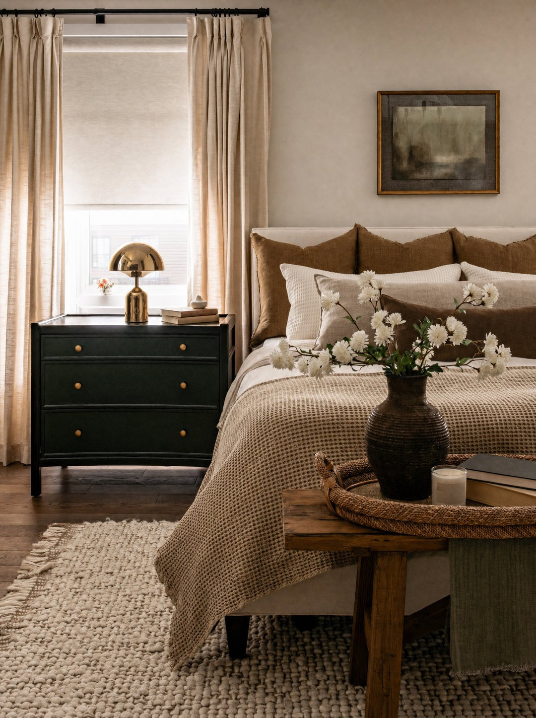 Vespers, a gold mushroom lamp lit on a dark blue dresser beside two books, in a small bedroom with linen bedding and a beige curtain