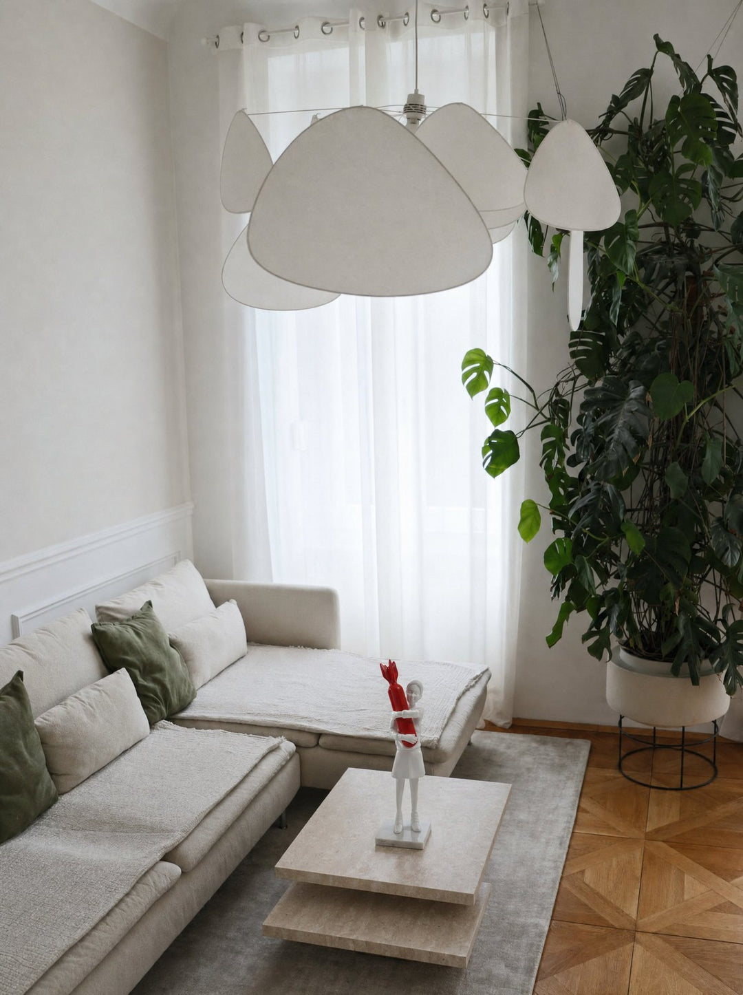 Disarm, a white figure holding a red bomb to its chest, on a low stone coffee table in front of a cream sofa, with sheer curtains and a monstera