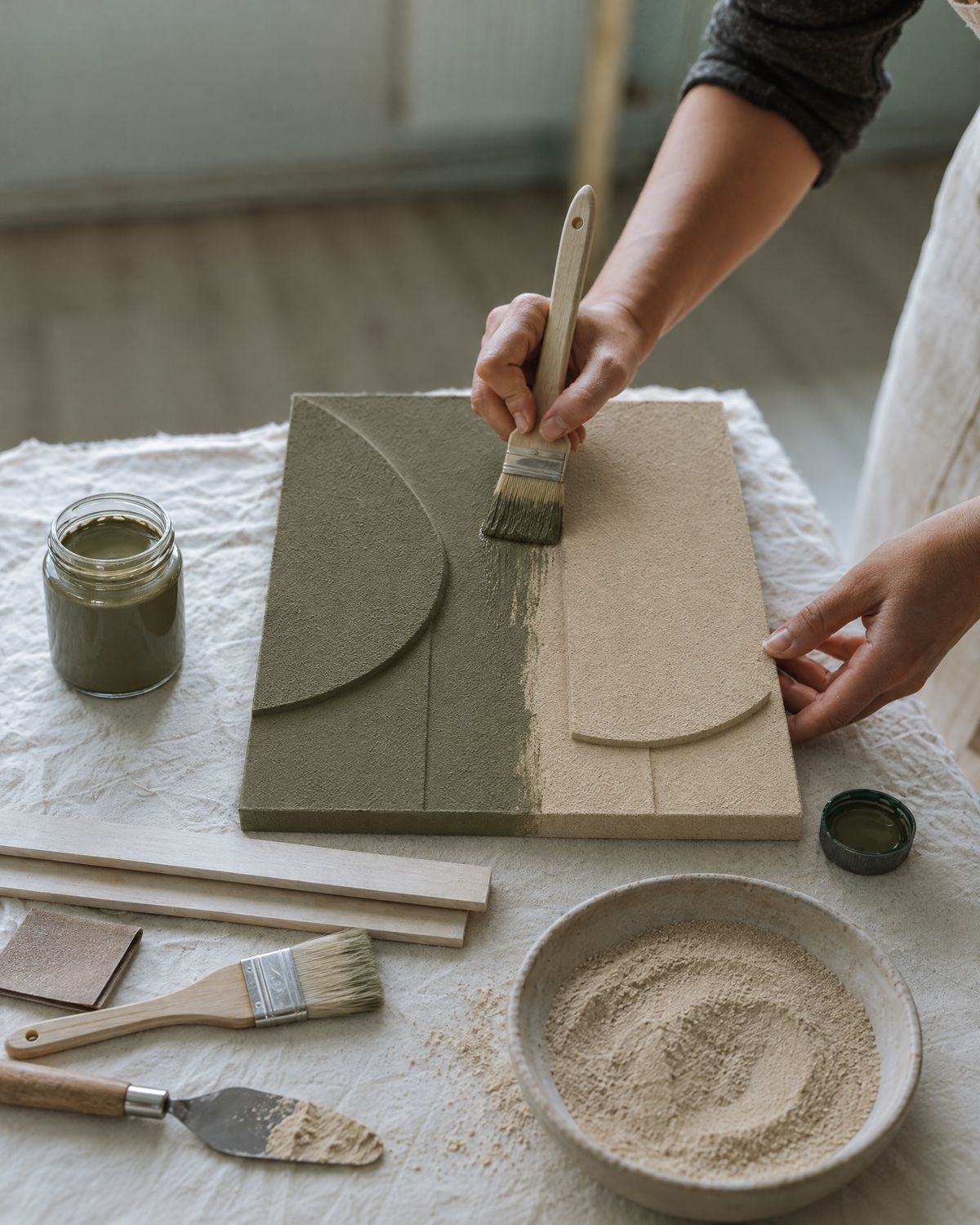 An artisan brushes Oliva, a grayed olive green, over the fine sand surface of an unframed Tinta panel, half done and half still beige, beside a jar of paint, a bowl of fine sand, a palette knife with sand paste, sandpaper and a spare brush